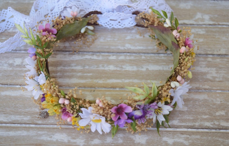 May include: A floral crown made of dried flowers and greenery. The crown is made of a variety of flowers, including white daisies, purple flowers, and pink flowers. The crown is on a wooden background.