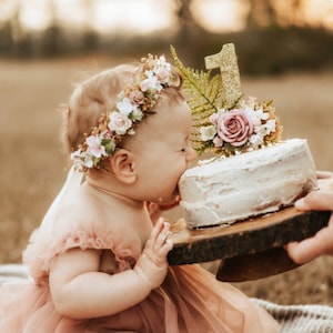 May include: A baby girl wearing a flower crown is about to take a bite out of a white frosted cake decorated with a gold glitter number one and pink flowers. The cake is on a wooden cake stand.