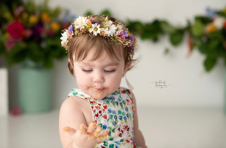 May include: A young child wearing a flower crown and a floral print dress is covered in cake frosting. The child is smiling and looking at the camera.