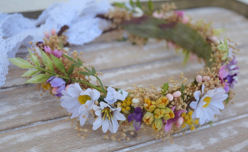 May include: A floral crown made of white, yellow, purple, and pink flowers. The crown is made of dried flowers and greenery.