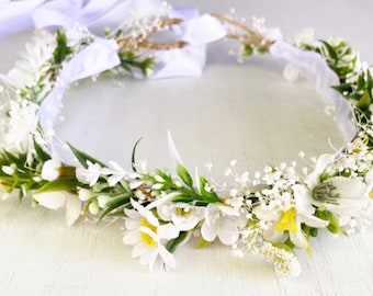 Könnte beinhalten: Eine weiße Blumenkrone mit kleinen weißen Blüten und grünen Blättern. Die Krone ist mit einem weißen Band und einer braunen Drahtbasis gefertigt.