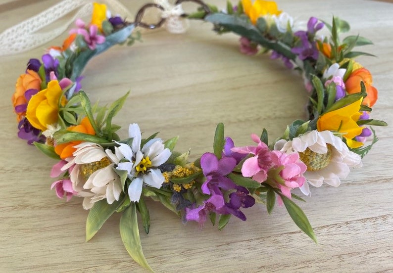 May include: A floral crown with yellow, orange, white, pink, and purple flowers. The crown is made with a wire base and is decorated with greenery.