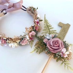 K&ouml;nnte beinhalten: Ein Blumenkranz mit rosa und wei&szlig;en Bl&uuml;ten und einem goldenen Glitzer-Nummer-Eins-Topper mit rosa und wei&szlig;en Bl&uuml;ten.