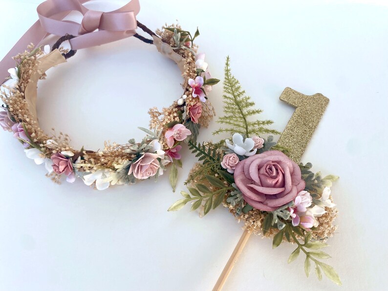 K&ouml;nnte beinhalten: Ein Blumenkranz mit rosa und wei&szlig;en Bl&uuml;ten und einem goldenen Glitzer-Nummer-Eins-Topper mit rosa und wei&szlig;en Bl&uuml;ten.