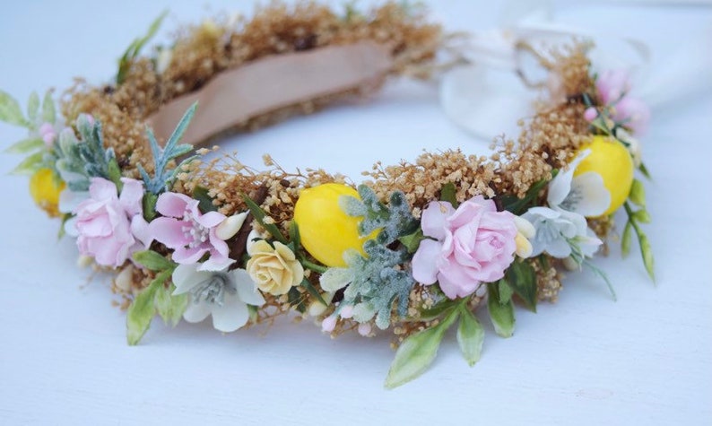 May include: A floral crown with pink and white flowers, green leaves, and yellow lemons. The crown is made of dried flowers and is perfect for a summer wedding or festival.