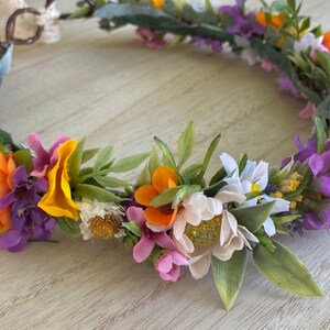May include: A floral crown with orange, yellow, white, pink, and purple flowers. The crown is made of artificial flowers and greenery.