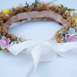 May include: A flower crown made of dried flowers and greenery with a white satin ribbon tied in a bow. The crown is decorated with small yellow and pink flowers.