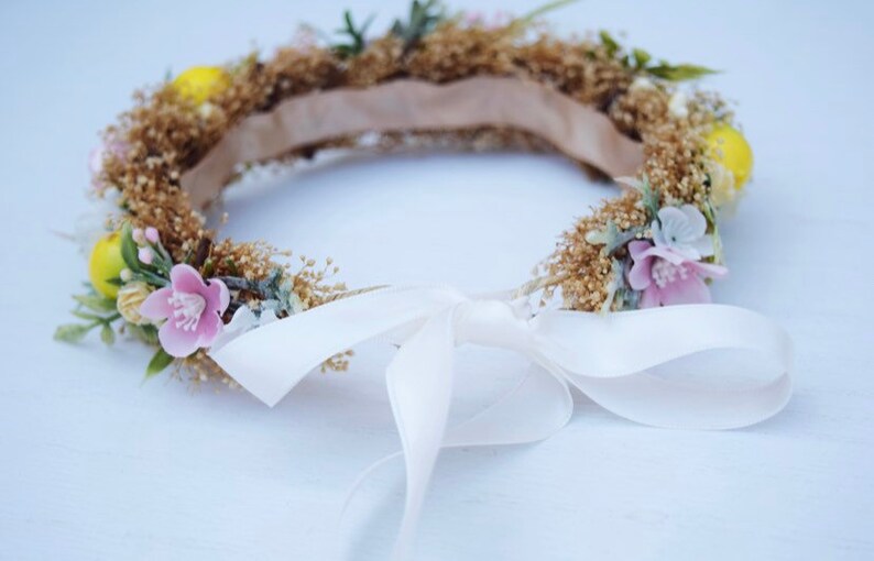 May include: A flower crown made of dried flowers and greenery with a white satin ribbon tied in a bow. The crown is decorated with small yellow and pink flowers.