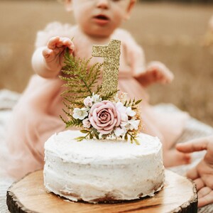 K&ouml;nnte beinhalten: Ein wei&szlig;er Kuchen mit einer goldenen Glitzer-Zahl eins als Topper und rosa Blumen auf einem Holzkuchenst&auml;nder. Ein junges M&auml;dchen in einem rosa Kleid und einer Blumenkrone greift nach dem Kuchen.