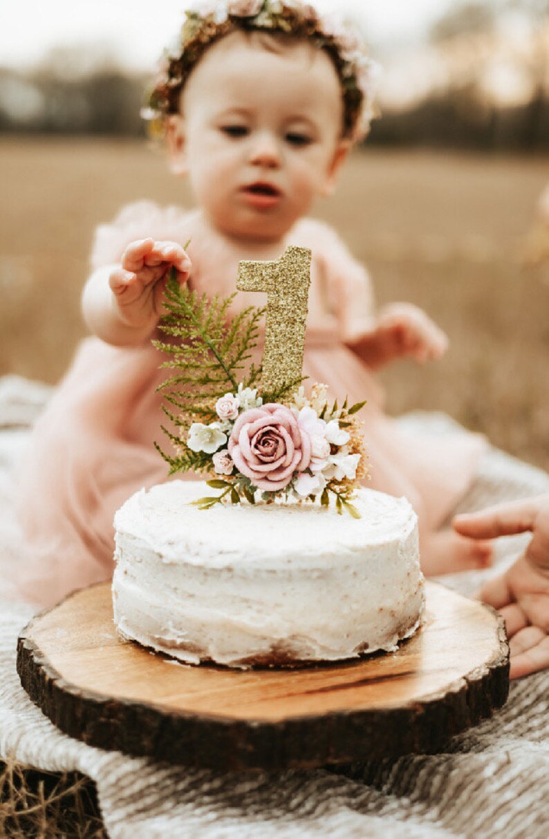 K&ouml;nnte beinhalten: Ein wei&szlig;er Kuchen mit einer goldenen Glitzer-Zahl eins als Topper und rosa Blumen auf einem Holzkuchenst&auml;nder. Ein junges M&auml;dchen in einem rosa Kleid und einer Blumenkrone greift nach dem Kuchen.