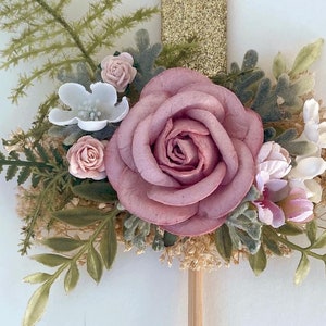 K&ouml;nnte beinhalten: Eine rosa Rose mit gr&uuml;nen Bl&auml;ttern und kleinen wei&szlig;en und rosa Bl&uuml;ten. Die Blumen sind an einem Holzstab befestigt.