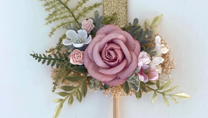 K&ouml;nnte beinhalten: Eine rosa Rose mit gr&uuml;nen Bl&auml;ttern und kleinen wei&szlig;en und rosa Bl&uuml;ten. Die Blumen sind an einem Holzstab befestigt.
