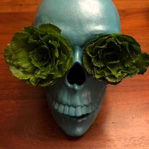 May include: A teal-colored skull with green floral accents covering the eye sockets. The skull has a metallic sheen and is set against a dark wooden surface. The skull's mouth is slightly open, revealing the teeth.