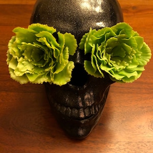 May include: A black skull with a glittery finish, adorned with two vibrant green, ruffled flower accents in place of eyes. The skull is set against a warm, wooden surface, creating a striking contrast.