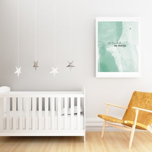 Op de afbeelding: Een wit babybedje met een wit matras en een groene aquarelprint met de tekst "for this child we prayed" hangt aan de muur erboven. Er staat een gele stoel in de hoek van de kamer.