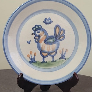 May include: A ceramic plate with a hand-painted rooster design. The plate features a blue border and a white center with a rooster in blue, orange, and green. The plate is displayed on a dark wooden stand.