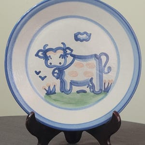 May include: A white ceramic plate with a hand-painted cow design. The cow is outlined in blue with pink spots and is set against a green field and blue sky. The plate has a blue border and is displayed on a wooden stand.