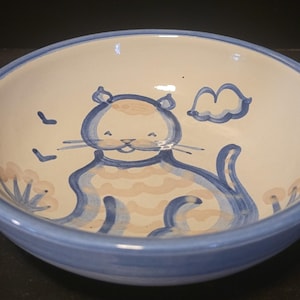 May include: A ceramic bowl with a light blue rim and a white interior. The bowl features a hand-painted design of a cat in blue and tan, with a cloud and birds. The bowl is suitable for serving food.