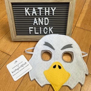 Könnte beinhalten: Eine Adler-Maske aus Filz mit gelbem Schnabel, grauen Augenbrauen und Augenlöchern. Die Maske ist weiß und hat ein Gummiband. Ein gerahmtes Buchstabenbrett zeigt "KATHY AND FLICK" in weißen Buchstaben. Ein kleines Etikett lautet "handmade just for you by kathyandflick".
