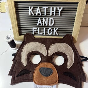 May include: A brown felt raccoon mask with white teeth, brown nose, and grey eyes. The mask is on a white surface with a letter board behind it that says "Kathy and Flick".