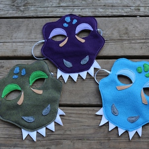 May include: Three felt dinosaur masks. One is purple, one is green, and one is blue. Each mask has a white felt teeth and eye details.