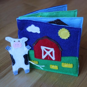 May include: A handmade felt farm-themed quiet book with a cow figure. The book features a red barn, a yellow sun, and a blue sky. The cow is white with black spots and a pink nose. The pages are various shades of blue and green.