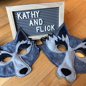 May include: Two gray and black felt wolf masks with black felt noses and eye holes. The masks are on a wooden floor with a gray letter board behind them that says "Kathy and Flick".