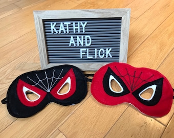 May include: Two felt Spider-Man masks, one black with a red spider web and one red with a black spider web. The masks are on a wooden surface with a letter board behind them that says "Kathy and Flick".