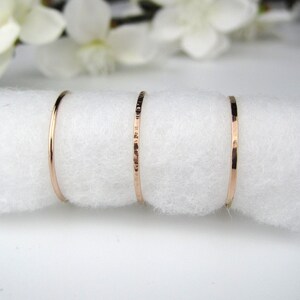May include: Four delicate rose gold rings displayed on a white, fluffy surface. The rings vary in design, with one smooth and three textured, showcasing a minimalist jewelry aesthetic. White flowers are softly blurred in the background.