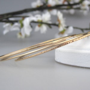 May include: Two gold-toned bangle bracelets, one with a hammered texture and the other smooth, are stacked on a white surface.