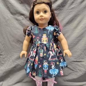 May include: A doll wearing a navy blue dress with a festive Nutcracker print, including ballerinas, toy soldiers, and Christmas trees. The doll has brown hair with pink bows and is wearing pink ballet shoes.