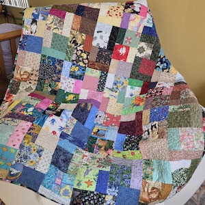 May include: A colorful patchwork quilt draped over a white chair. The quilt features a variety of fabric squares in numerous colors and patterns, including floral, animal, and geometric designs. The quilt's edges are slightly curved, and it appears to be handmade.