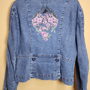 May include: A blue denim jacket with a floral embroidery on the back. The embroidery features pink flowers and green leaves. The jacket has a classic collar, button closures, and two decorative buttons at the waist.