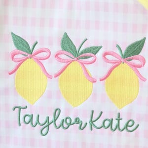 May include: Embroidered design featuring three yellow lemons with green leaves and pink bows on a pink and white gingham background. The name "Taylor Kate" is written in green cursive below the lemons.
