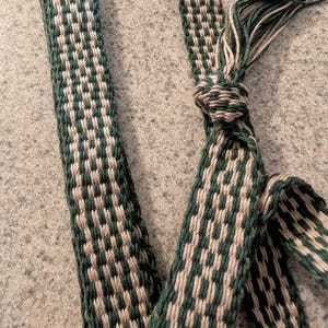 Könnte beinhalten: Nahaufnahme eines gewebten Gürtels oder Riemens in einem sich wiederholenden Muster aus Grün und Off-White. Der Riemen ist an einem Ende verknotet, mit losen Fäden. Der Stoff hat ein strukturiertes Aussehen.