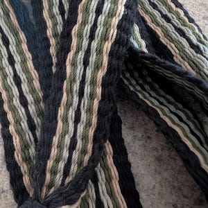 Puede incluir: Primer plano de un textil tejido con un patrón repetitivo de rayas negras, verde oliva y beige. El textil parece estar hecho de hilo o cuerda gruesa, con una superficie texturizada. El diseño es una mezcla de colores oscuros y neutros.