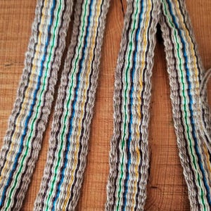 May include: A woven textile strap featuring a repeating pattern of beige, dark brown, bright blue, green, and yellow vertical stripes. The fabric has a textured, corded appearance with visible interlaced threads throughout the entire length.