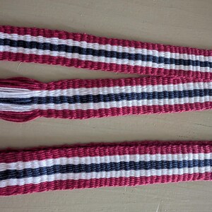 May include: A woven fabric belt or strap featuring a repeating striped pattern in deep red, white, and dark navy blue. The textile has a textured, ribbed appearance and includes a decorative tassel end with loose white, navy, and red threads.