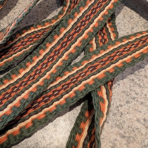 Puede incluir: Primer plano de una cinta textil tejida con un patrón geométrico repetitivo. La cinta presenta una paleta de colores verde, naranja, crema y marrón. El diseño incluye rayas verticales y elementos en forma de diamante.
