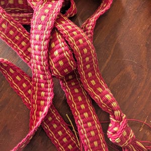 Puede incluir: Primer plano de un textil tejido en tonos rojos y amarillos. El textil presenta un patrón repetitivo de cuadrados amarillos sobre un fondo rojo. El material tejido está anudado en un extremo, con hilos sueltos visibles.