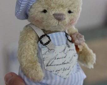 May include: A small, cream-colored plush teddy bear wearing a light blue and white striped beret and matching overalls. The overalls feature a white fabric patch on the chest with cursive black script text. The bear has dark eyes and a stitched nose.