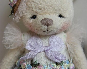 May include: A handmade teddy bear with a cream-colored body, wearing a light purple dress and a straw hat adorned with flowers. The bear has a lavender bow and a floral belt. The bear's face has rosy cheeks and a gentle expression.
