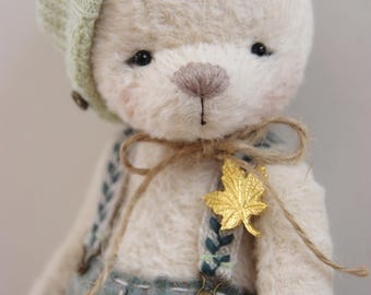 May include: A cream-colored plush teddy bear wearing a light green knit beret and blue-green plaid trousers with suspenders. The bear features a brown stitched nose, pink-tinted cheeks, and a gold-colored maple leaf pin attached to the twine bow.