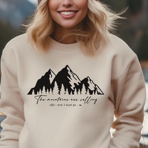 Könnte beinhalten: Ein beiger Pullover mit einem schwarzen Bergketten-Motiv und dem Text "The mountains are calling - and I must go -"