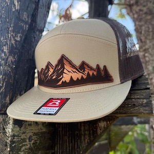 May include: A tan flat bill snapback hat with a brown mesh back and a mountain graphic. The graphic features brown and orange mountains and trees. The hat has a white tag that reads "Flat Bill Snapback".