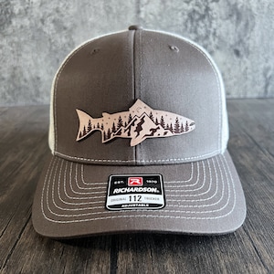 May include: A brown and white trucker hat with a brown leather patch featuring a silhouette of a fish with mountains and trees in the background. The patch also has a small moon and stars. The hat has the text "Richardson. Original 112 Trucker Adjustable" on the back.