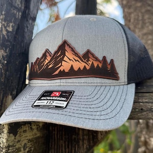 May include: Gray trucker hat with a mountain and forest design. The hat features a brown and orange mountain graphic on the front panel, a gray brim, and a black mesh back. The label reads "RICHARDSON ORIGINAL 112 TRUCKER ADJUSTABLE."