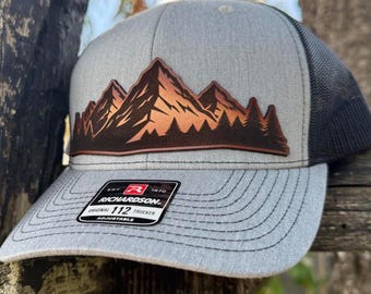May include: Grey trucker hat with a mountain and forest design. The hat has a brown and orange mountain graphic on the front panel, a grey brim, and a black mesh back. The label reads "RICHARDSON ORIGINAL 112 TRUCKER ADJUSTABLE."