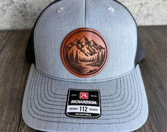 May include: A grey trucker cap with a black mesh back and a brown leather patch depicting a mountain and forest scene. The cap has the text "RICHARDSON ORIGINAL 112 TRUCKER ADJUSTABLE".