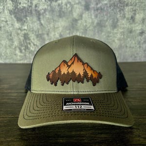 Może przedstawiać: Zielona i czarna czapka trucker z haftowanym pasmem górskim i drzewami z przodu. Czapka jest regulowana i ma napis "Richardson. Original 112 Trucker Adjustable" z tyłu.